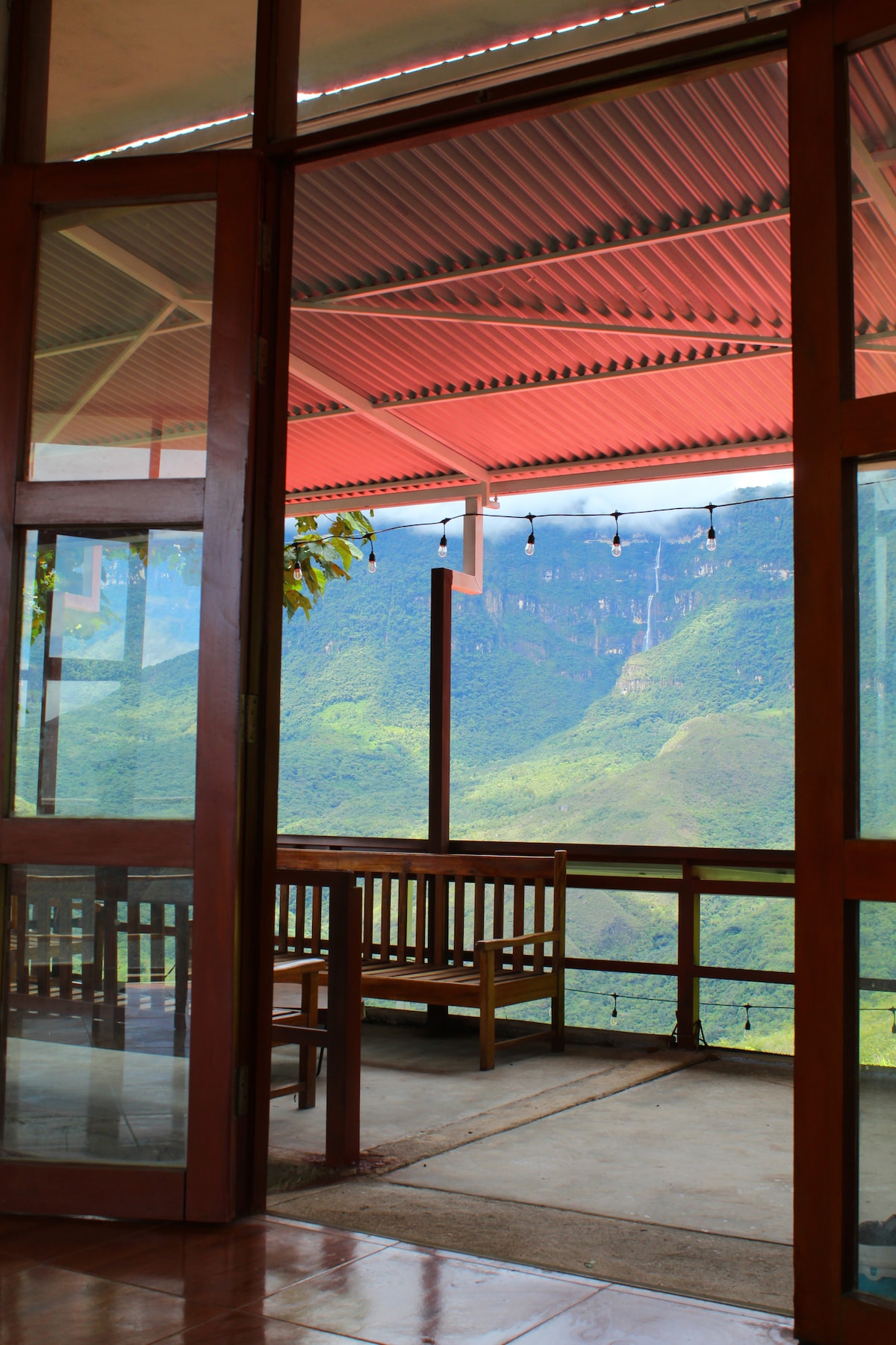 A spacious terrace is visible through large glass doors, offering a view of lush green mountains and valleys. Wooden seating is arranged on the terrace, beneath a red roof. Soft outdoor lighting hangs above, enhancing the inviting space for relaxation.
