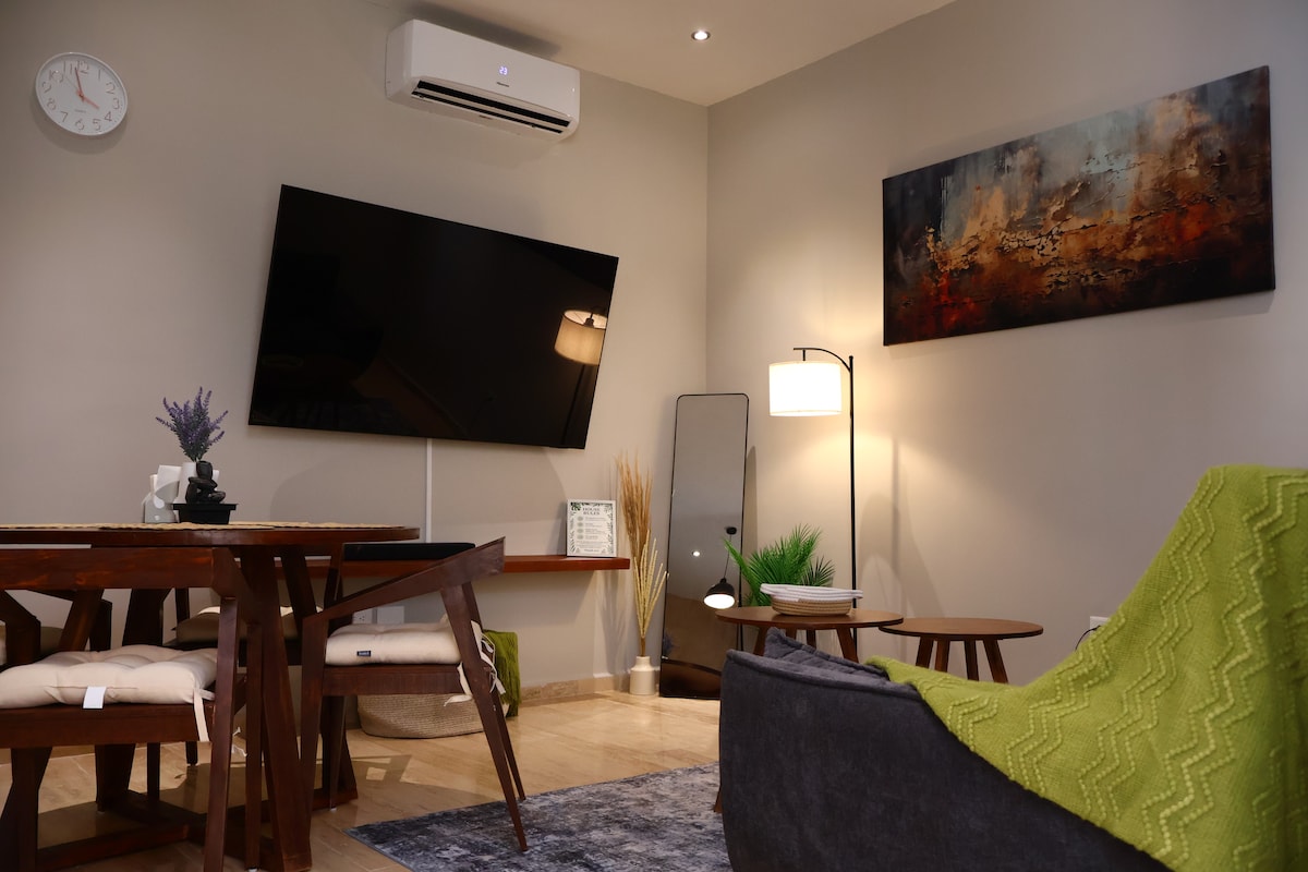 A modern living space is depicted with a large wall-mounted television and a stylish dining table. A comfortable seating area features a plush sofa with a textured throw. Soft lighting is provided by a standing lamp, and a mirror adds depth to the room.