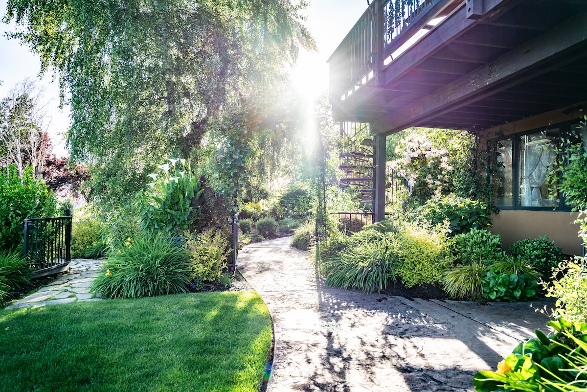 A well-maintained garden path is framed by lush greenery and vibrant flowers, leading towards a staircase under a covered porch. Gentle sunlight filters through the leaves, creating a serene atmosphere. The stone pathway curves gracefully, inviting guests to explore the beautifully landscaped surroundings.