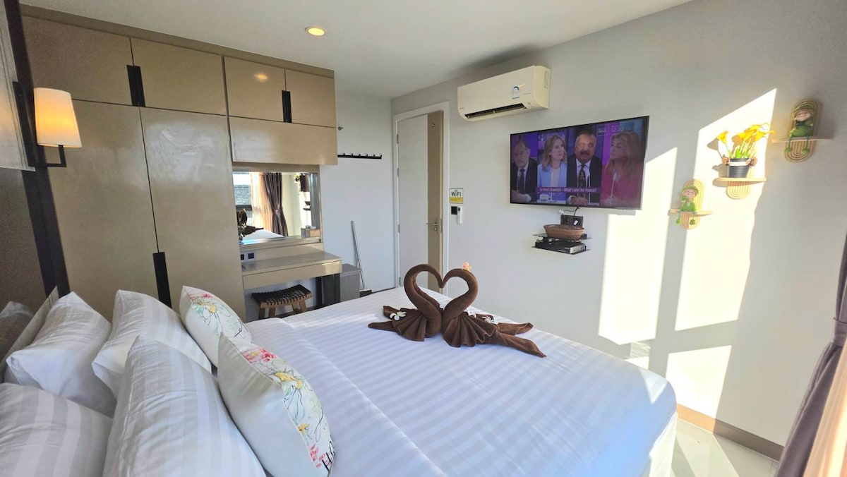 A cozy bedroom is presented with a spacious bed adorned with decorative pillows. A heart-shaped towel arrangement is placed at the center. A wall-mounted TV is visible, alongside modern cabinetry. Natural light fills the room, creating a comfortable atmosphere.