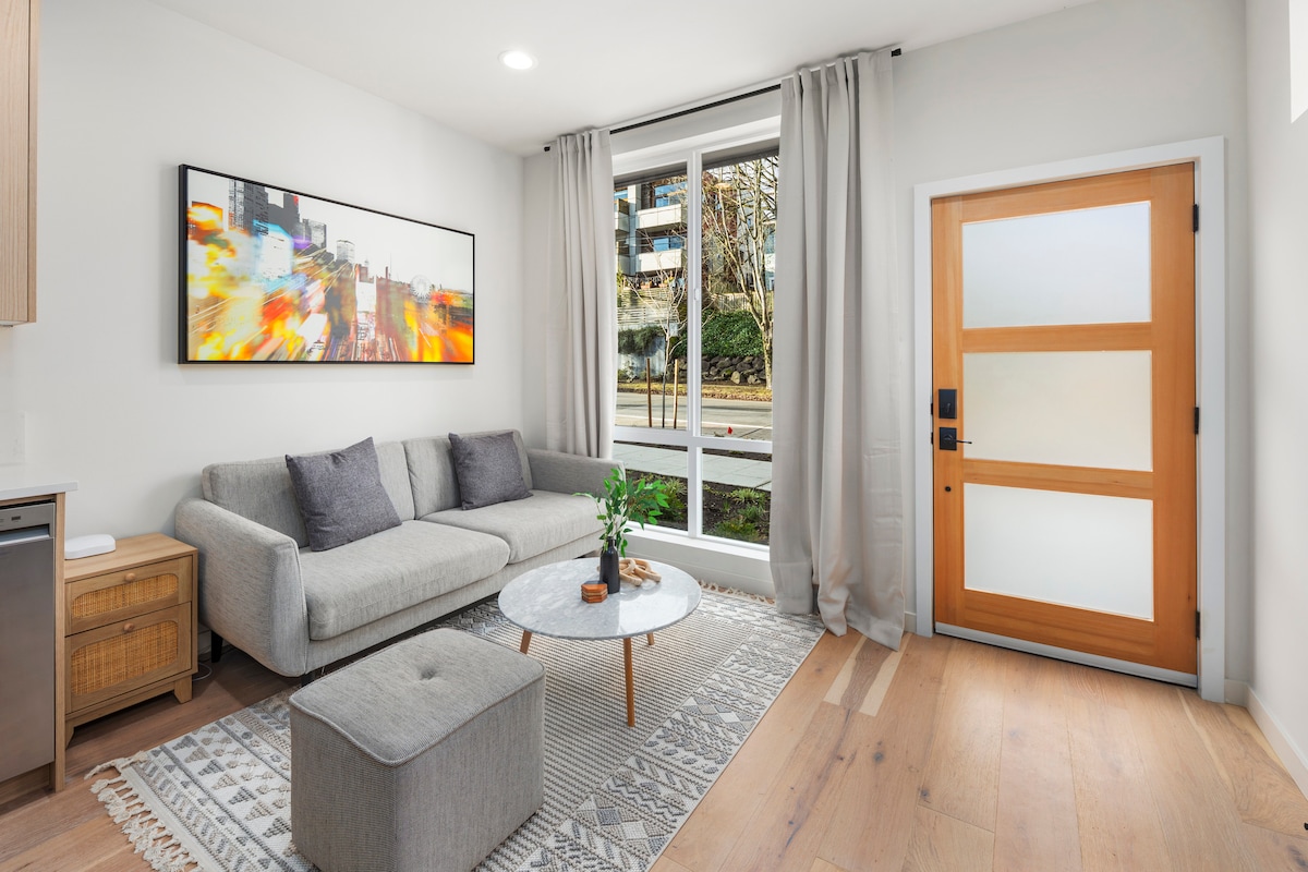 A modern living area features a light grey sectional sofa paired with a soft ottoman. A round coffee table sits atop a patterned area rug, and a large artwork adds a splash of color on the wall. Natural light pours in through the window, creating a bright atmosphere.