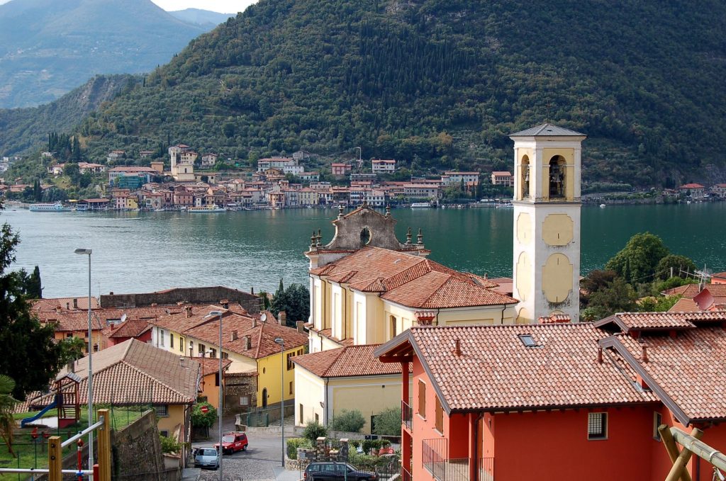 A scenic view of a lakeside town is captured, featuring terracotta-roofed buildings with a bell tower prominently in the foreground. The expansive lake reflects the surrounding hills, with greenery framing the picturesque village in the distance.