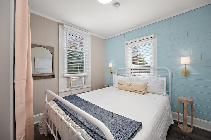 Serene bedroom with a soft blue wall, bright windows, and fresh white bedding perfect for unwinding and sleeping well