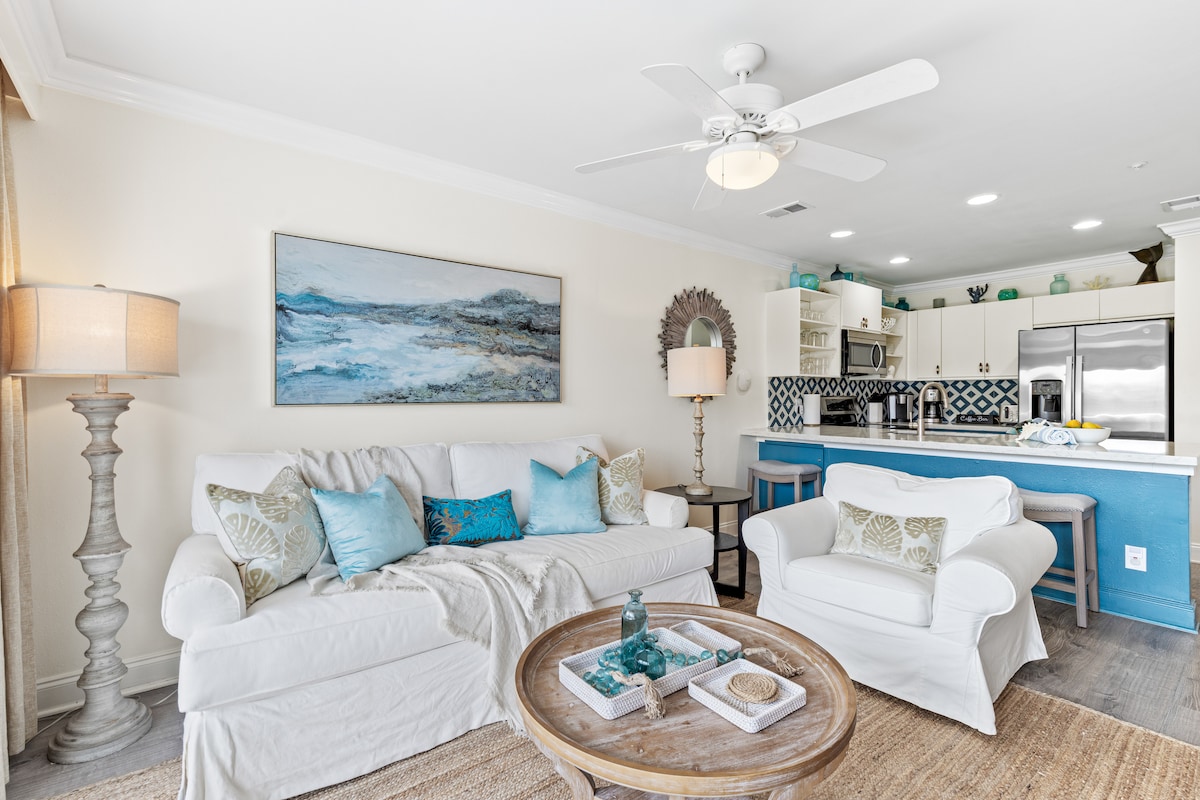 The living area features a white sofa adorned with blue and green accent pillows. A round wooden coffee table sits at the center, complementing the light wood flooring. A ceiling fan and decorative wall art enhance the space, while the kitchen area is visible in the background.