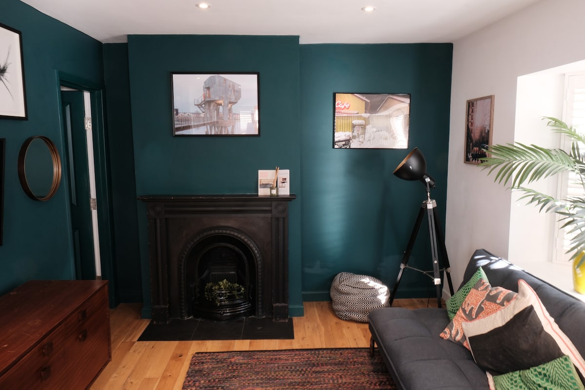 A cozy lounge area features a dark teal accent wall, complemented by framed artwork and a decorative mirror. A modern floor lamp stands near the sofa, and a patterned throw adorns the seating. A Victorian-style fireplace adds character, with a small plant positioned on the hearth.