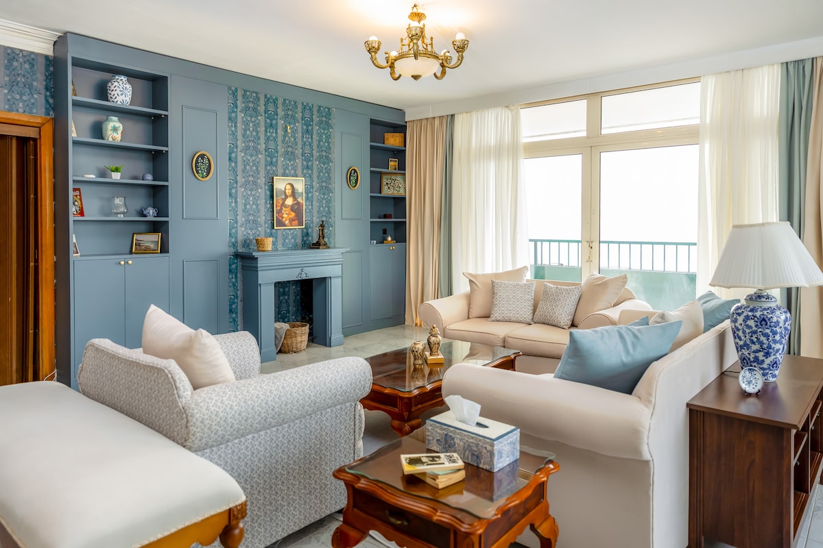 A bright living area features comfortable seating arrangements with plush sofas and a vintage coffee table. Shelves display decorative items against a soft blue accent wall. Large windows invite natural light, creating a welcoming ambiance. A small balcony is visible through the glass doors.
