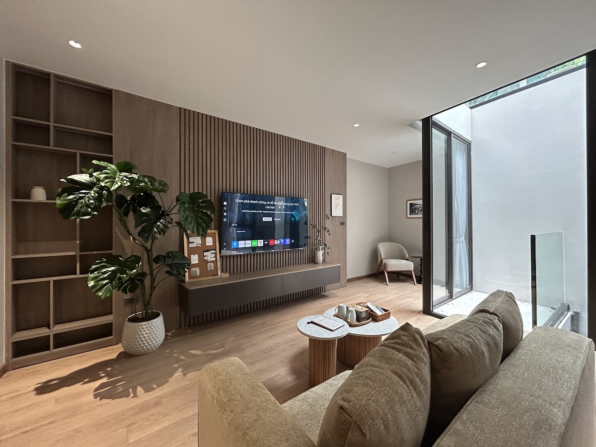 A spacious living area is highlighted by a large flat-screen TV mounted on a wood-paneled wall. A comfortable sofa is paired with a round coffee table. Natural light is filtered through large glass doors that open to an outdoor area, complemented by a decorative plant.