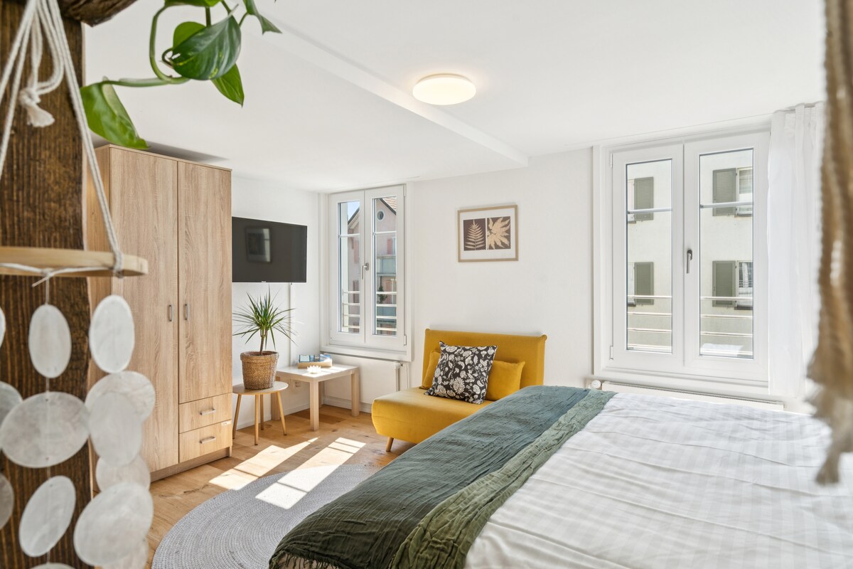 A bright room features a spacious bed adorned with a textured throw, complemented by a cozy armchair in yellow. A modern television is mounted on light-colored cabinetry, while natural light filters through two windows. A small potted plant adds a touch of greenery.