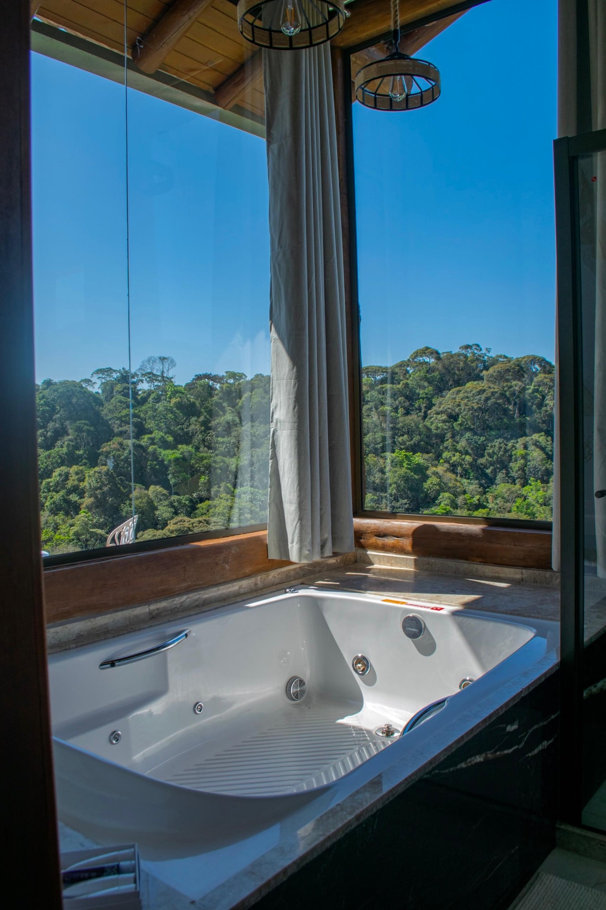 A bright bathroom features a spacious bathtub positioned by a large window, offering panoramic views of lush greenery. Natural light floods the area, highlighting the clean lines of the tub and the elegant curtain draping beside it.