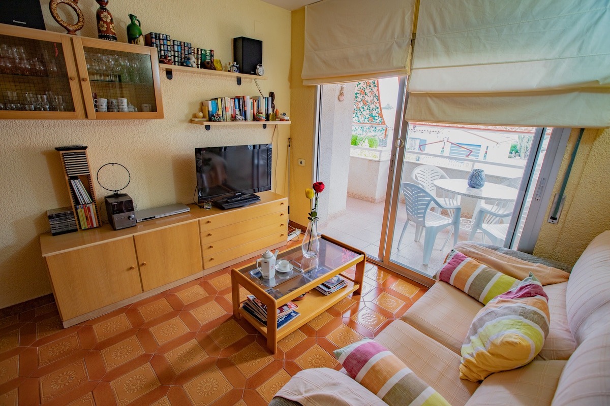 A cozy living area features a comfortable sofa with colorful cushions and a glass coffee table. A television is positioned on a wooden cabinet, complemented by shelves displaying books and decorative items. Natural light floods the space through sliding glass doors, leading to a balcony with outdoor seating.