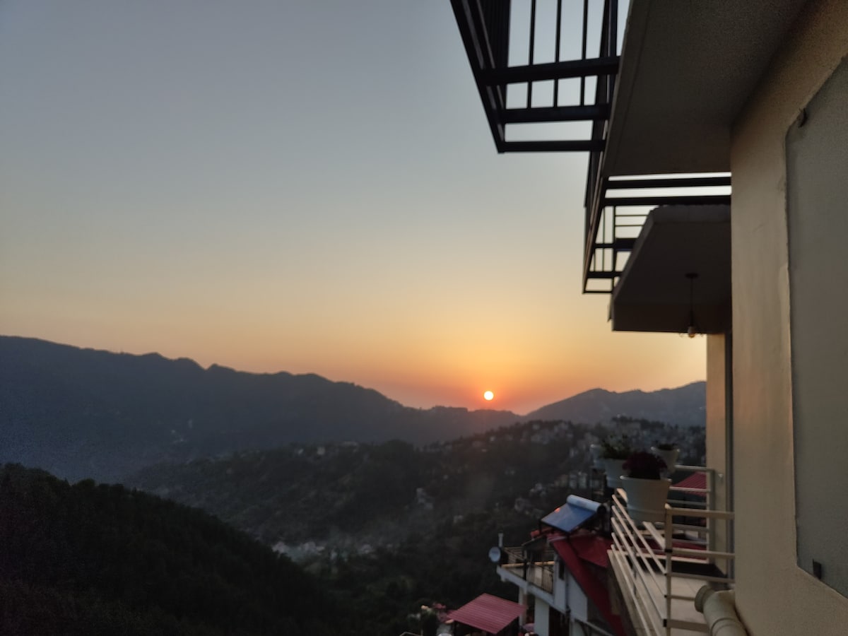 A breathtaking sunset is captured over the distant mountains, with the sun descending towards the horizon. The image features a balcony railing and potted plants that frame the view, enhancing the scenic beauty of the surrounding landscape.