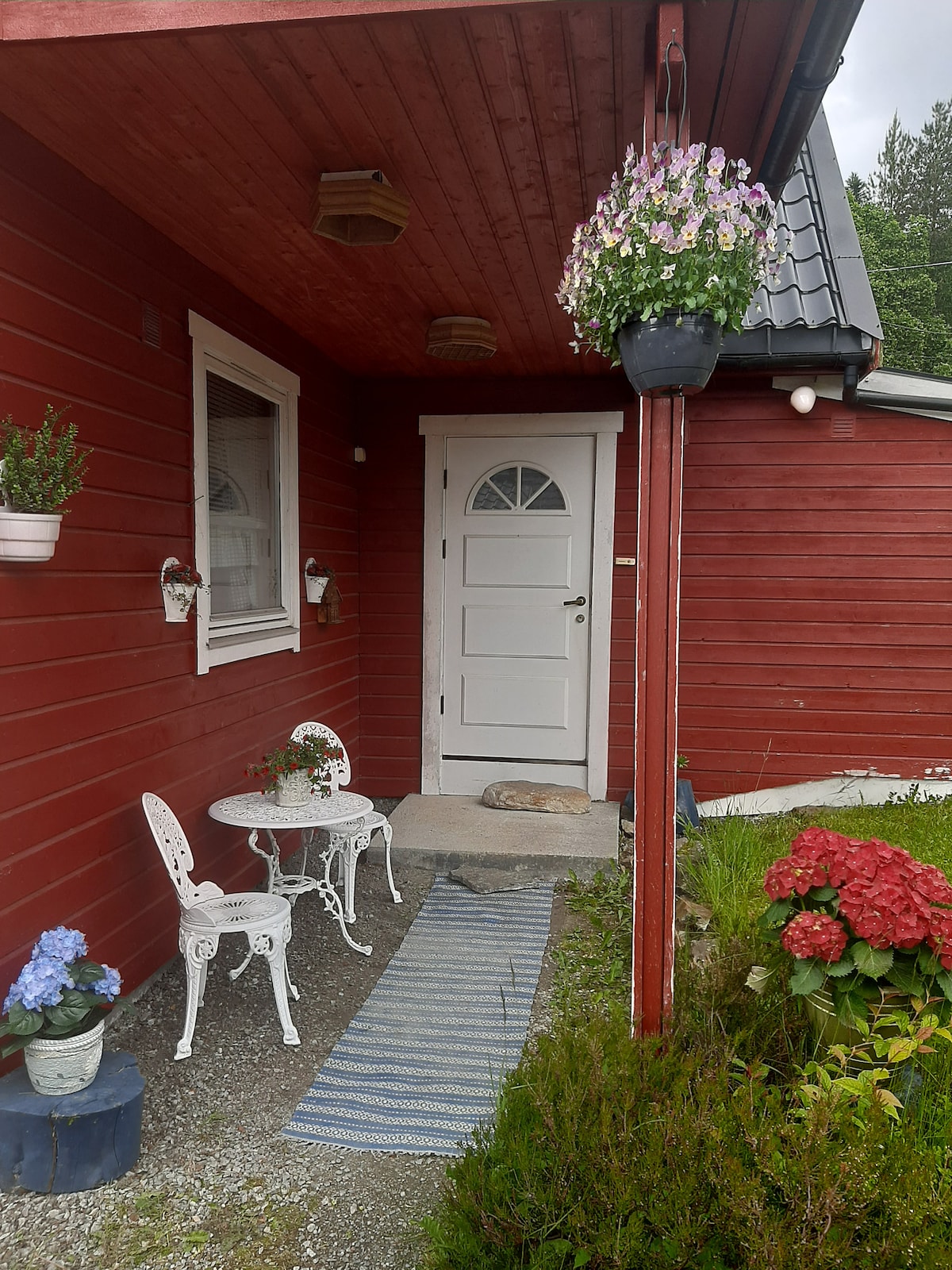 The entrance features a charming red wooden facade, where a white door stands framed by two small windows. A small table and two white chairs are positioned on a stone path, accompanied by potted flowers and a welcome mat. A striped blue rug adds texture to the entrance.