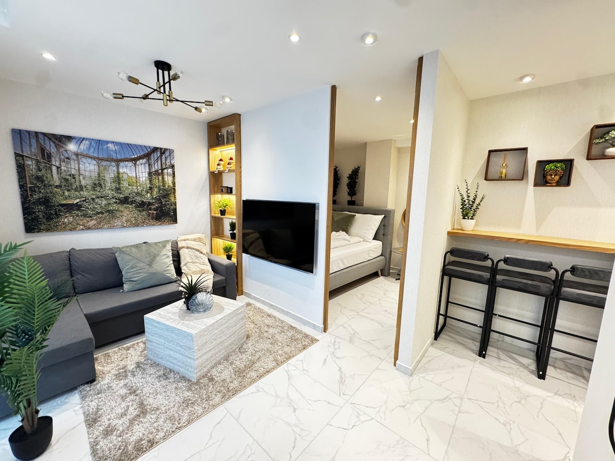 A modern living area features a gray sectional sofa positioned near a textured coffee table. Soft lighting highlights a large artwork depicting a forest scene. A compact kitchen space with bar stools is visible, along with a partial view of the bedroom sectioned with a subtle divider.