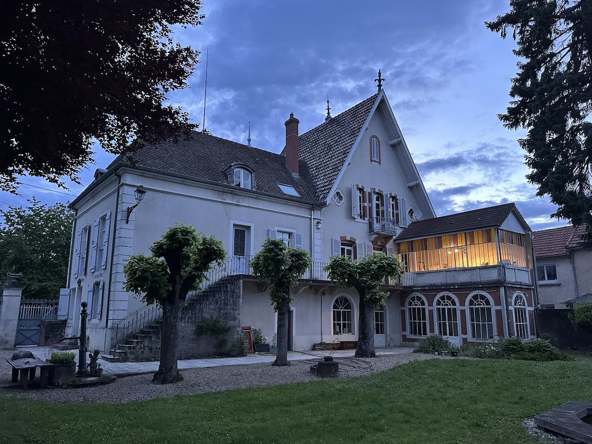 A grand house is presented with a distinct architectural style, featuring a mix of classic and modern elements. Brightly lit windows illuminate the upper section as twilight descends, while well-maintained gardens and symmetrical trees form a serene outdoor space.