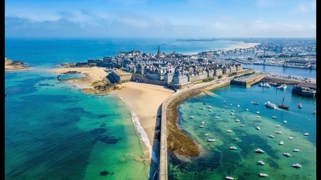 An aerial view shows the coastline of Saint-Malo, featuring historic fortifications along the sandy beaches and a vibrant blue sea. Small boats are anchored in the clear waters, and the town's distinctive architecture is visible against the contrasting natural landscape.