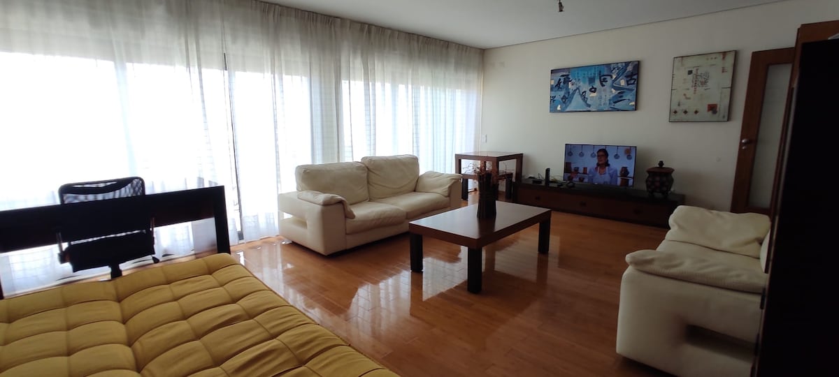 An airy living room is presented with large windows allowing ample natural light. A spacious brown coffee table occupies the center. Light-colored sofas are positioned across from a TV on a wooden stand, enhancing the welcoming feel of the room.
