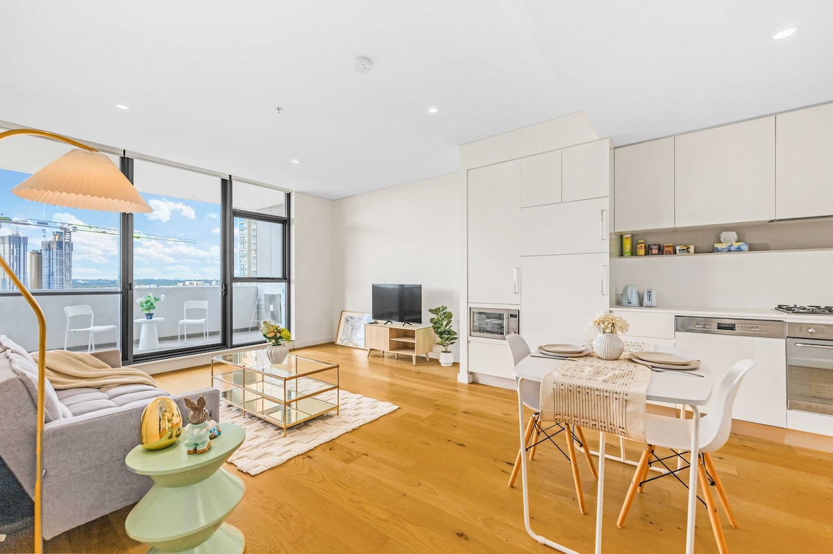 A bright living area is shown with large windows offering panoramic views. A modern gray sofa sits adjacent to a glass coffee table. A smart TV is mounted on the wall, and a dining table with white chairs is visible beside a fully equipped kitchen with sleek cabinetry and appliances.