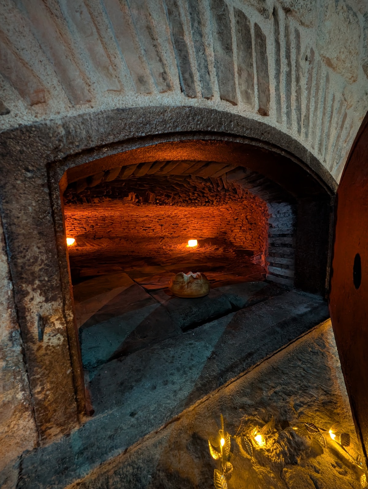 An interior view of a traditional bread oven is captured, featuring rustic stone walls illuminated by warm candlelight. The oven's arched entrance frames a glimpse of the oven's interior, where a round loaf sits on a surface, suggesting a sense of heritage and craftsmanship.