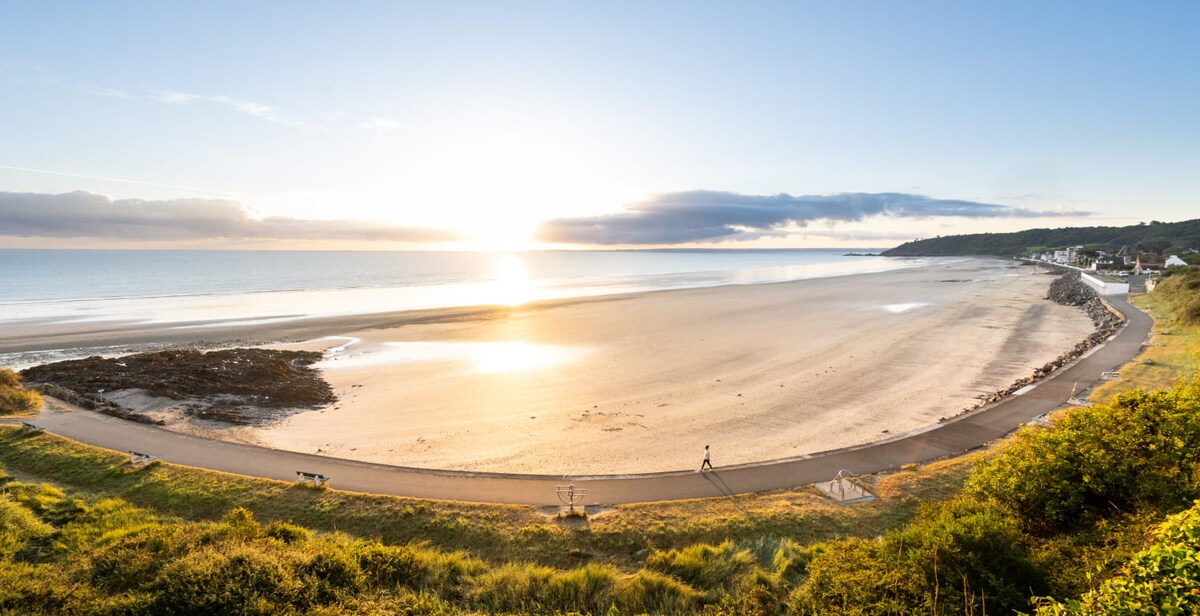 A serene beach scene is captured at sunset, with soft waves lapping at the sandy shore. A winding path leads along the beach, bordered by lush greenery. The horizon is filled with gentle hues of orange and blue, creating a calming atmosphere over the landscape.