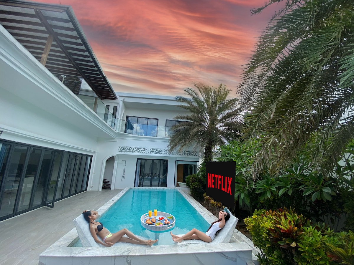 Two sun loungers are positioned in a private pool, with a small table holding colorful drinks and snacks. The surrounding foliage adds greenery, while a large outdoor screen displays a 'NETFLIX' logo. The sky is filled with vibrant sunset colors, enhancing the serene atmosphere.