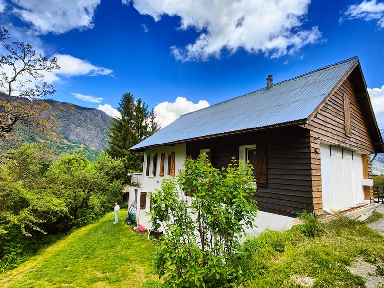The exterior of a spacious house is showcased against a backdrop of mountains and a vibrant blue sky. Lush greenery surrounds the property, with a well-maintained lawn. The house features a combination of wooden and stucco elements, with several windows and a sloping metal roof.