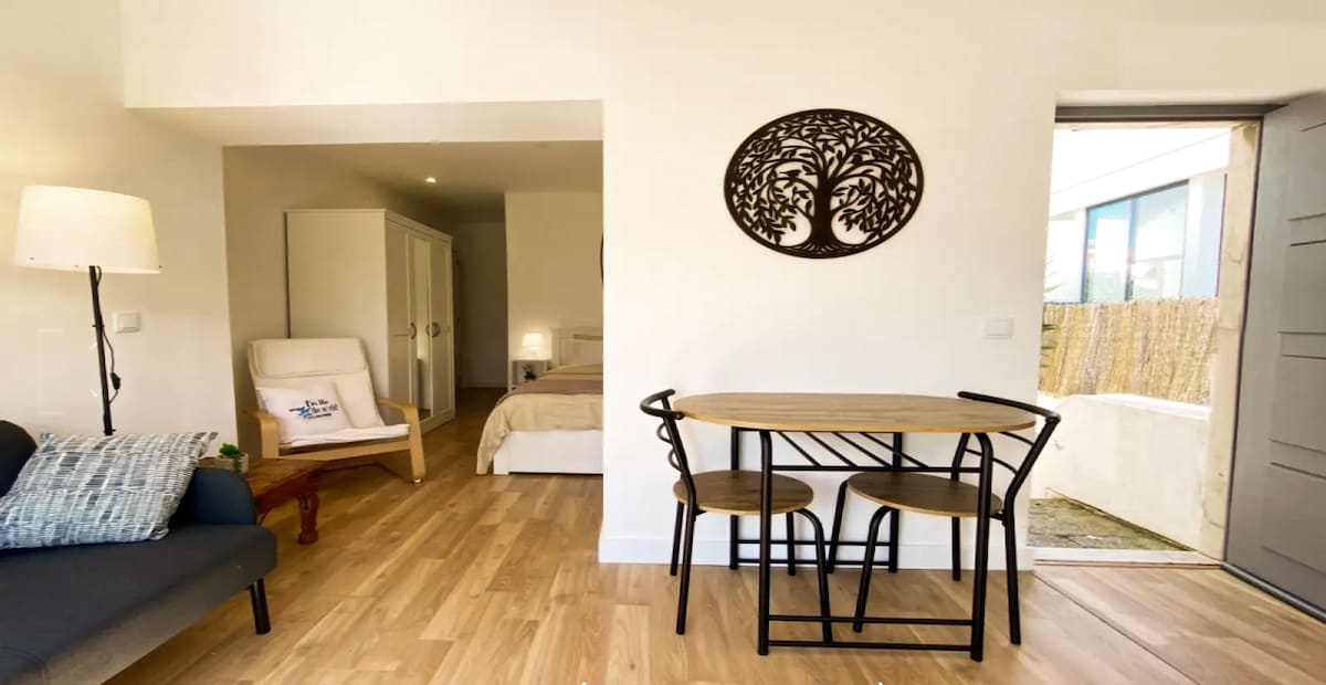 A bright and airy living space features a small dining table surrounded by two chairs. A decorative tree wall art enhances the atmosphere. A light-colored armchair and a sofa provide comfortable seating, and a bedroom area is visible through an open doorway.