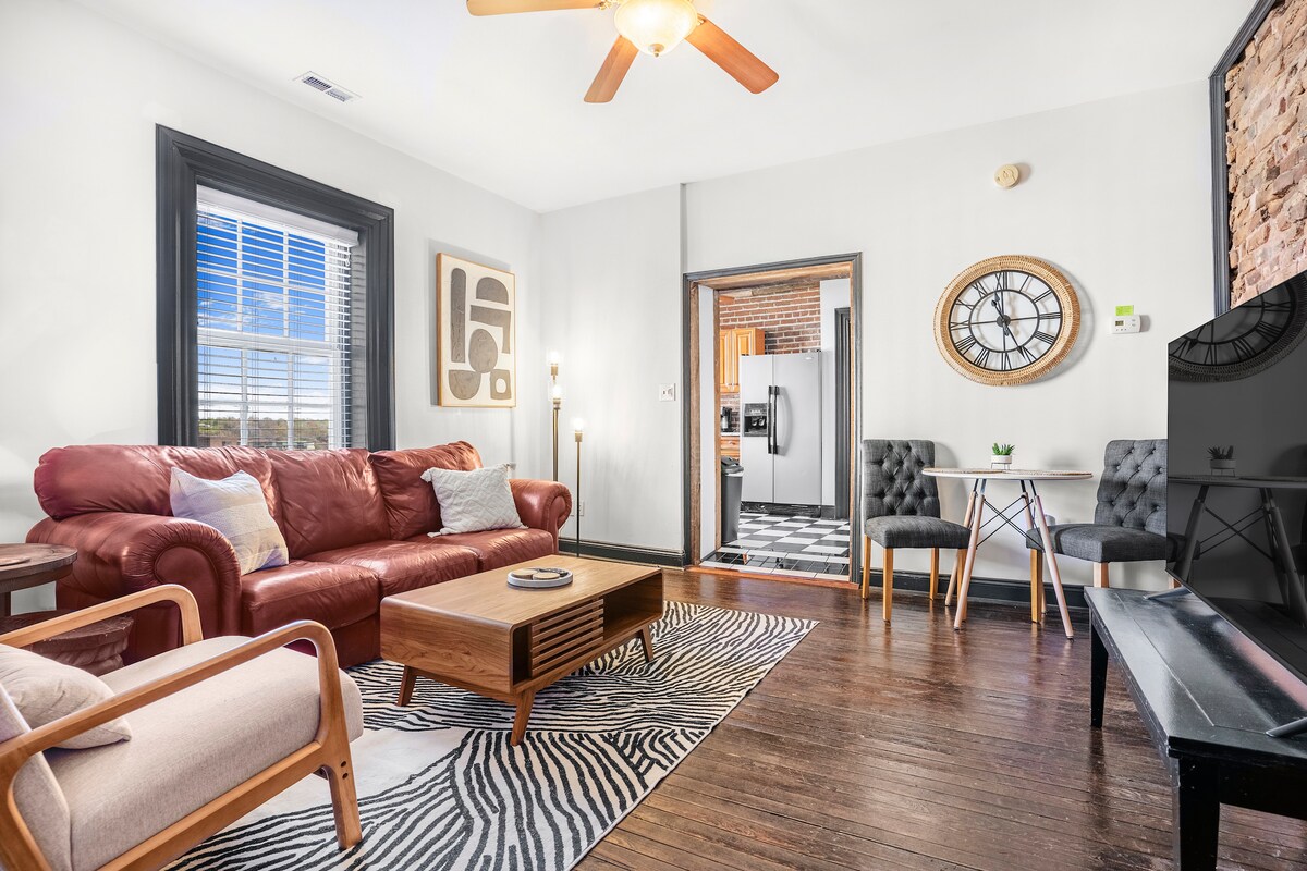 The living room features a cozy leather couch and two chairs, arranged around a wooden coffee table. A large smart TV sits opposite the seating area. Bright light filters through a large window, highlighting the hardwood floor and exposed brick wall in the background.