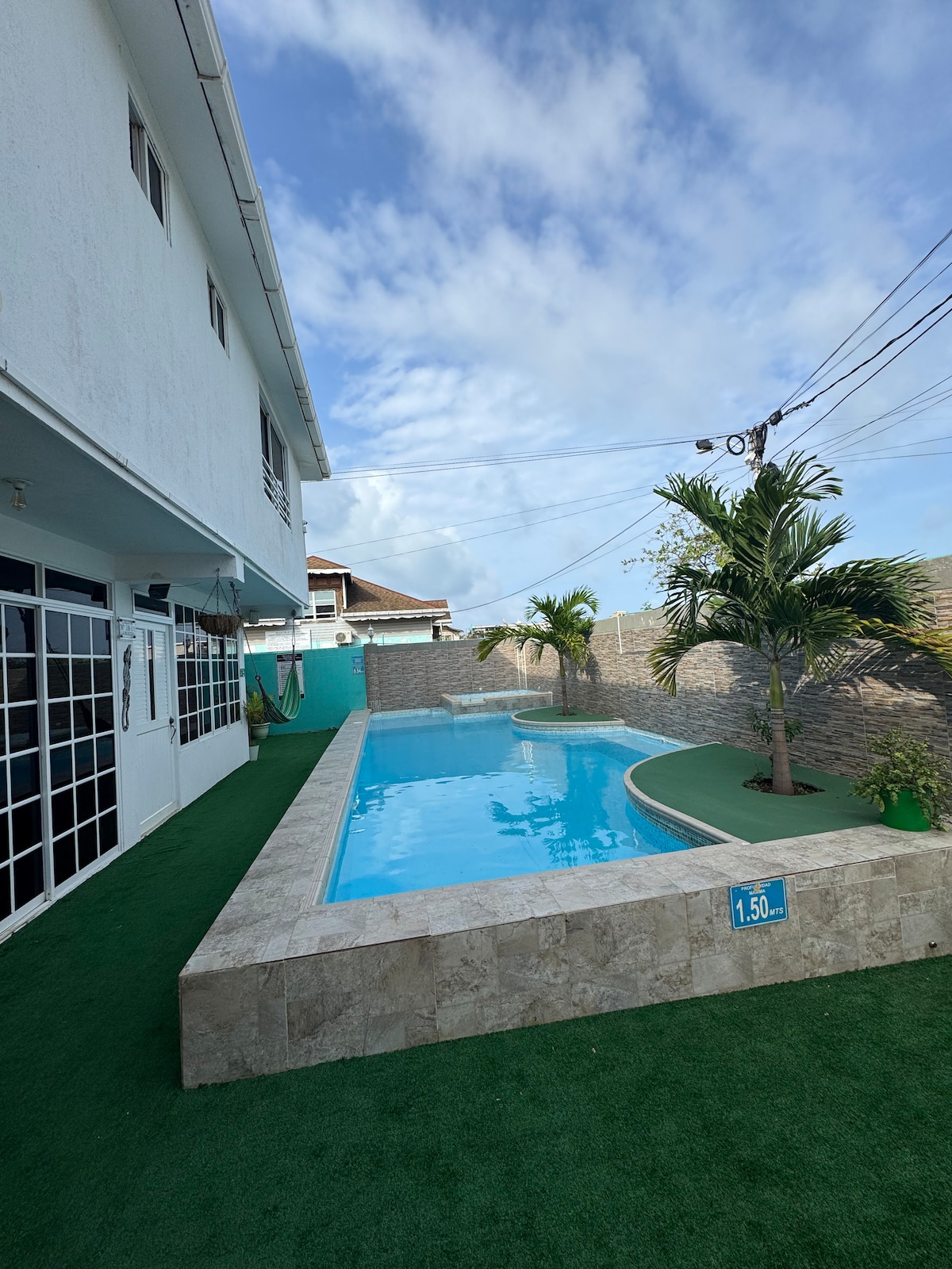 A serene outdoor area features a swimming pool surrounded by a stone edge and manicured greenery. The space is bordered by a textured wall, providing privacy. Clear blue skies and a few clouds are visible above, while the two-story house is partially seen in the background.