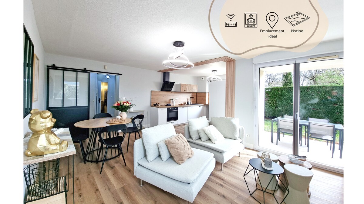 An open-plan living area features a modern kitchen and dining space. A circular dining table accommodates four people, while a cozy seating arrangement includes light-colored sofas. Large glass doors lead to an outdoor terrace, offering views of the greenery outside.