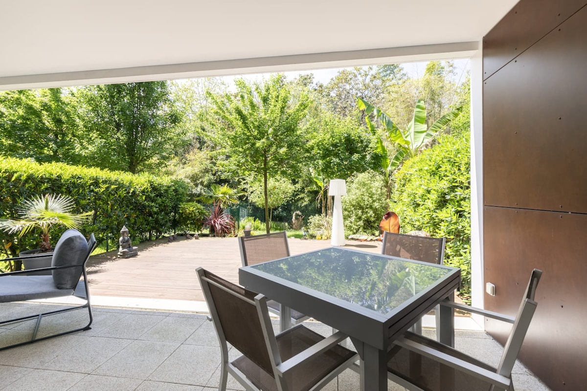 An outdoor terrace is presented with a modern dining table surrounded by four chairs. Lush greenery and trees enhance the view, creating a serene atmosphere. A lounging chair is positioned beside the table, facilitating relaxation in the private garden space.