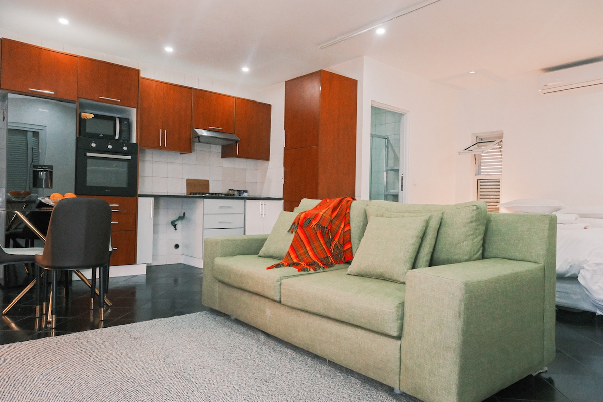 A compact studio apartment features a light green sofa with a colorful throw draped over it. A dining table is set nearby, accompanied by black chairs. The kitchen showcases wooden cabinetry and modern appliances, while a cozy sleeping area with a bed is visible in the background.