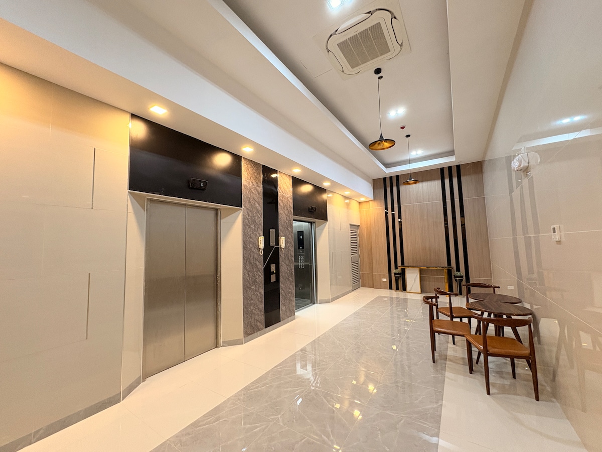 A modern lobby area showcases sleek surfaces and neutral tones. Elevators are situated centrally, framed by stylish lighting. A small seating area is arranged with wooden chairs around a round table, creating a welcoming space with ample natural light from large windows.