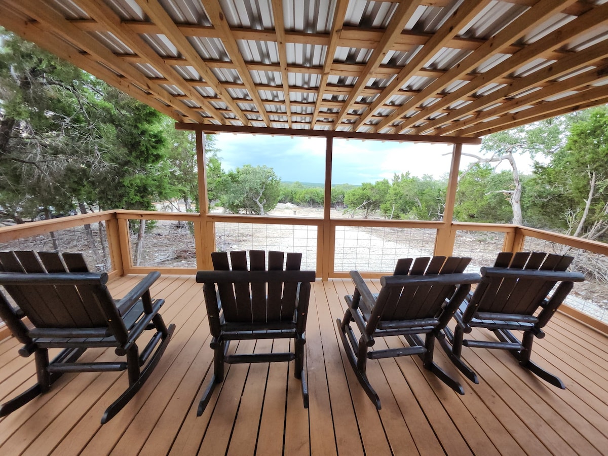 A spacious porch is seen with four wooden rocking chairs positioned facing an expansive view of the landscape. The ceiling is supported by wooden beams, providing a shaded area. A natural setting with greenery is visible beyond the porch.