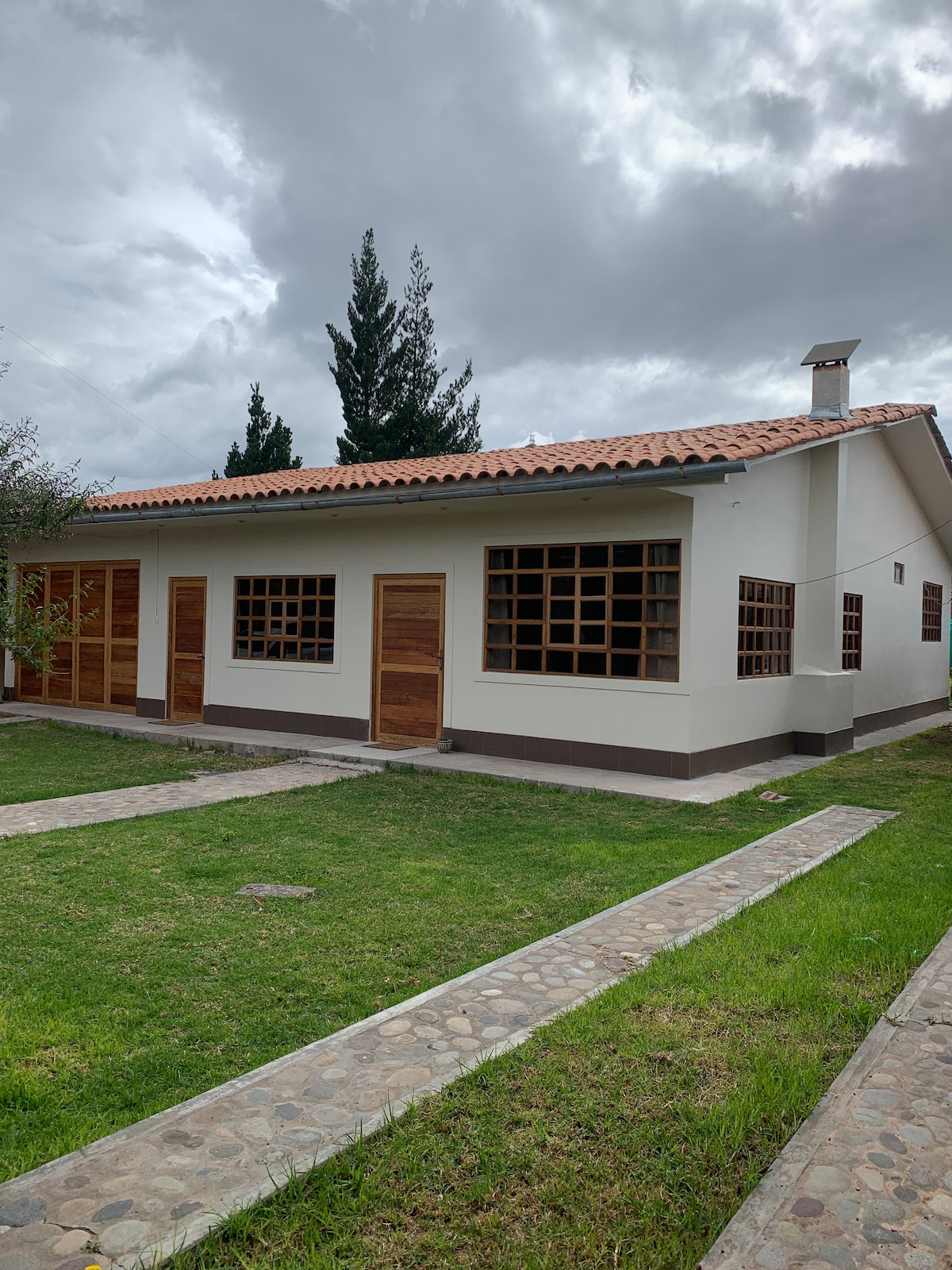 The exterior of a single-story house is depicted, featuring a light-colored façade with wooden doors and window frames. A well-maintained grassy area surrounds the structure. Gravel pathways lead to the entrance, with trees visible in the background under a cloudy sky.