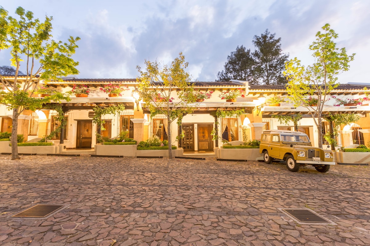 The exterior features a charming row of colonial-style villas, each with wooden doors and colorful flower arrangements. A yellow vintage vehicle is parked on the stone-paved pathway, which is bordered by lush greenery. Soft evening light casts a warm glow over the scene.