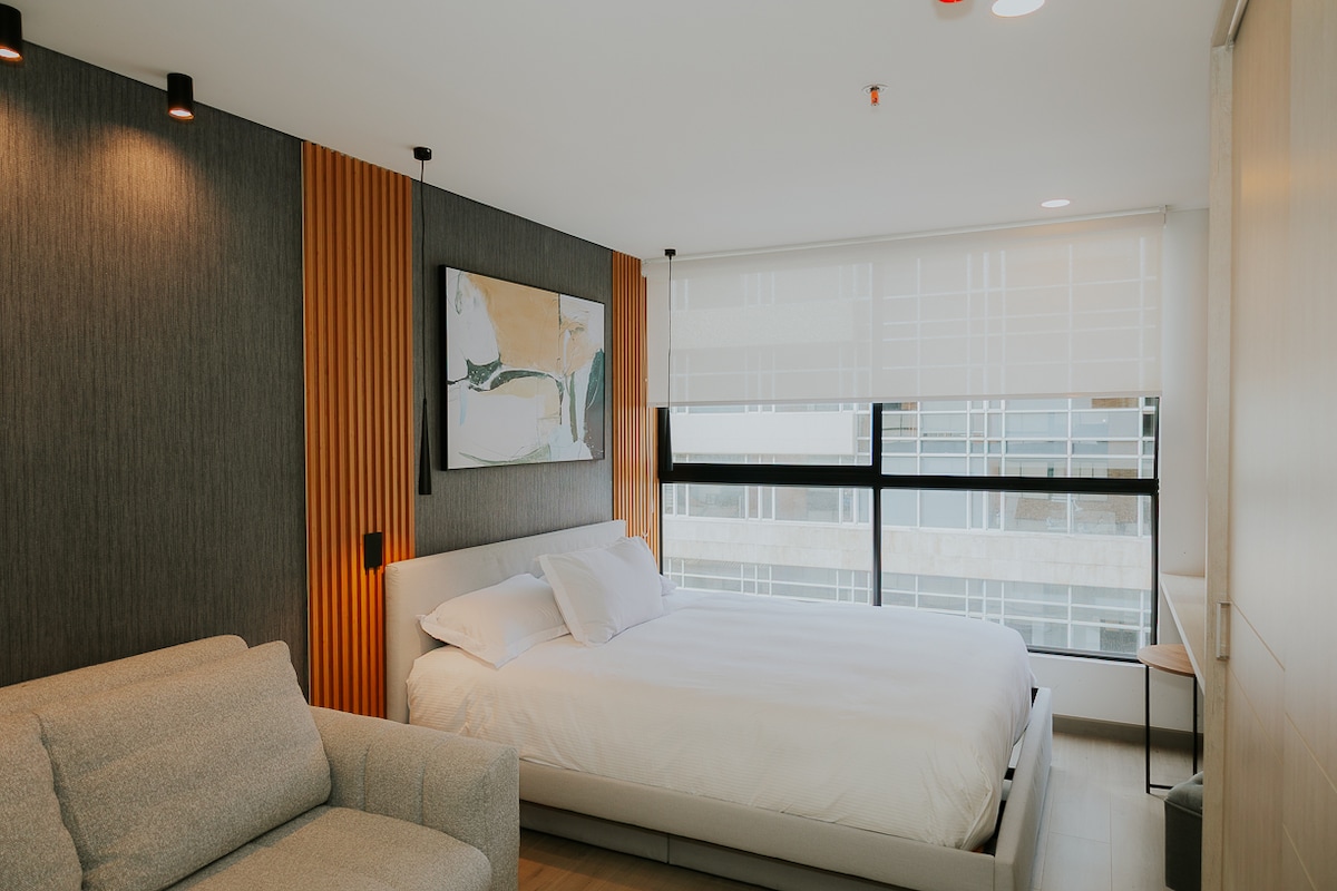 A modern bedroom features a spacious bed with crisp white linens. Natural light floods in through a large window dressed in sheer shades. A comfortable sofa sits beside the bed, with textured wall panels adding warmth to the space. A neutral color palette creates a serene environment.