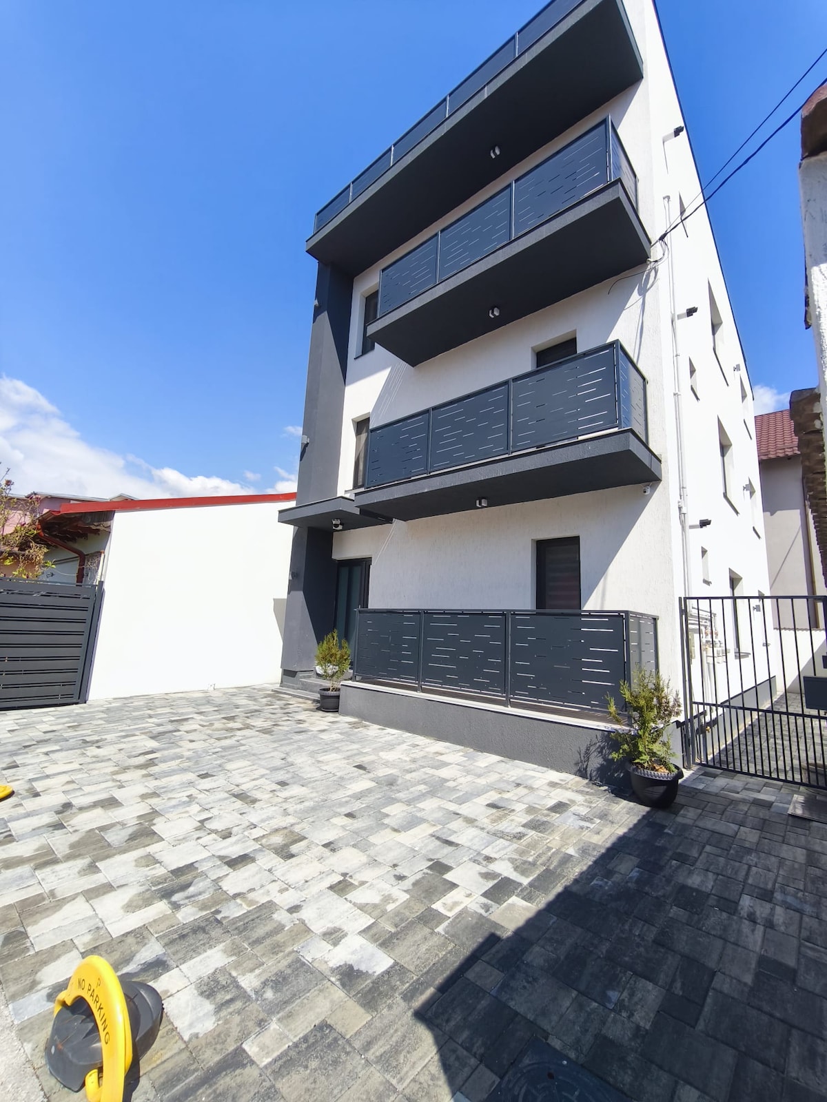 A modern building features three levels with balconies adorned by stylish railings. The façade showcases a clean white exterior contrasted by dark accents. A paved outdoor area with planters adds to the entrance's appeal, while a clear blue sky provides a bright backdrop.