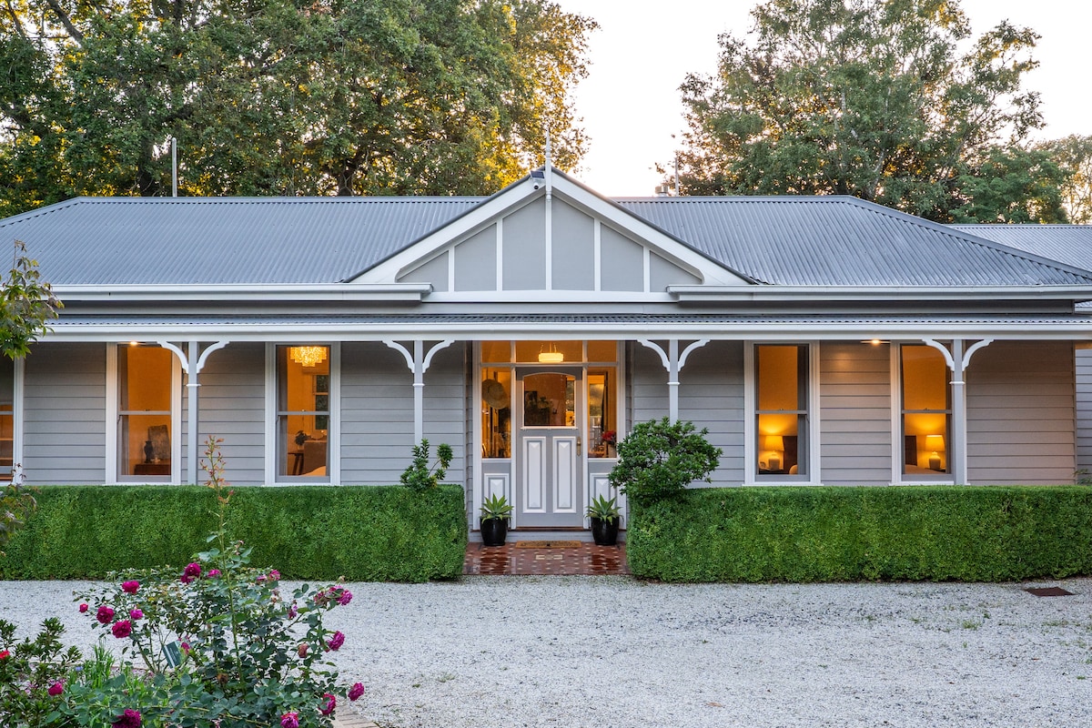 A charming cottage exterior is framed by neatly trimmed hedges and vibrant rose bushes. The traditional architecture features a welcoming entrance with a double door, complemented by large windows that softly illuminate the interior, reflecting a warm ambiance in the evening light.