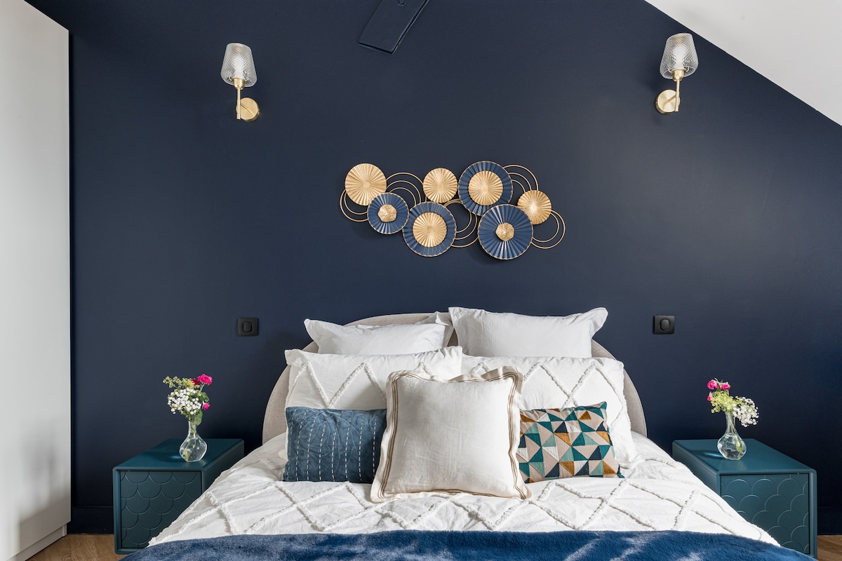 A spacious bed is adorned with a variety of decorative cushions, set against a dark blue wall. Two bedside tables hold small vases with fresh flowers, and wall sconces add soft lighting to the room's ambiance.