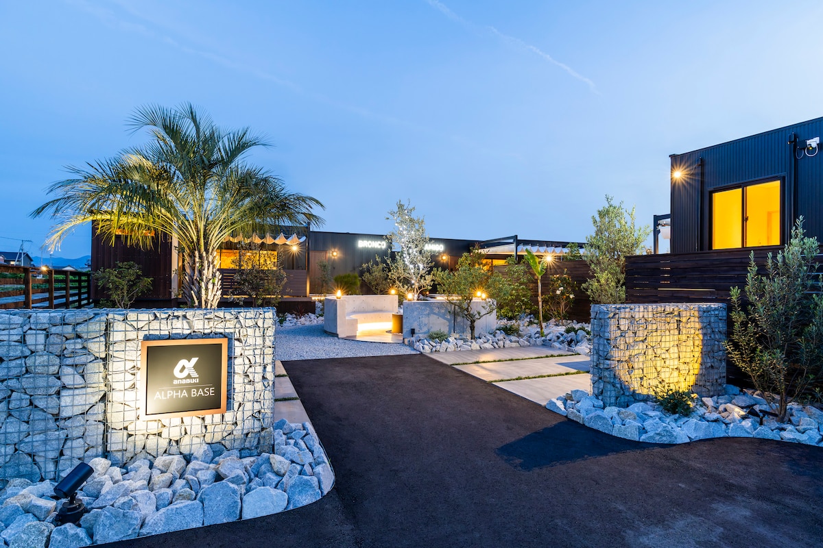 The entrance to ALPHA BASE is framed by landscaped gardens and stone pathways. The logo is illuminated on a decorative stone monument, enhancing the welcoming ambiance. A spacious parking area is visible, surrounded by greenery and soft lighting, creating a serene arrival experience.