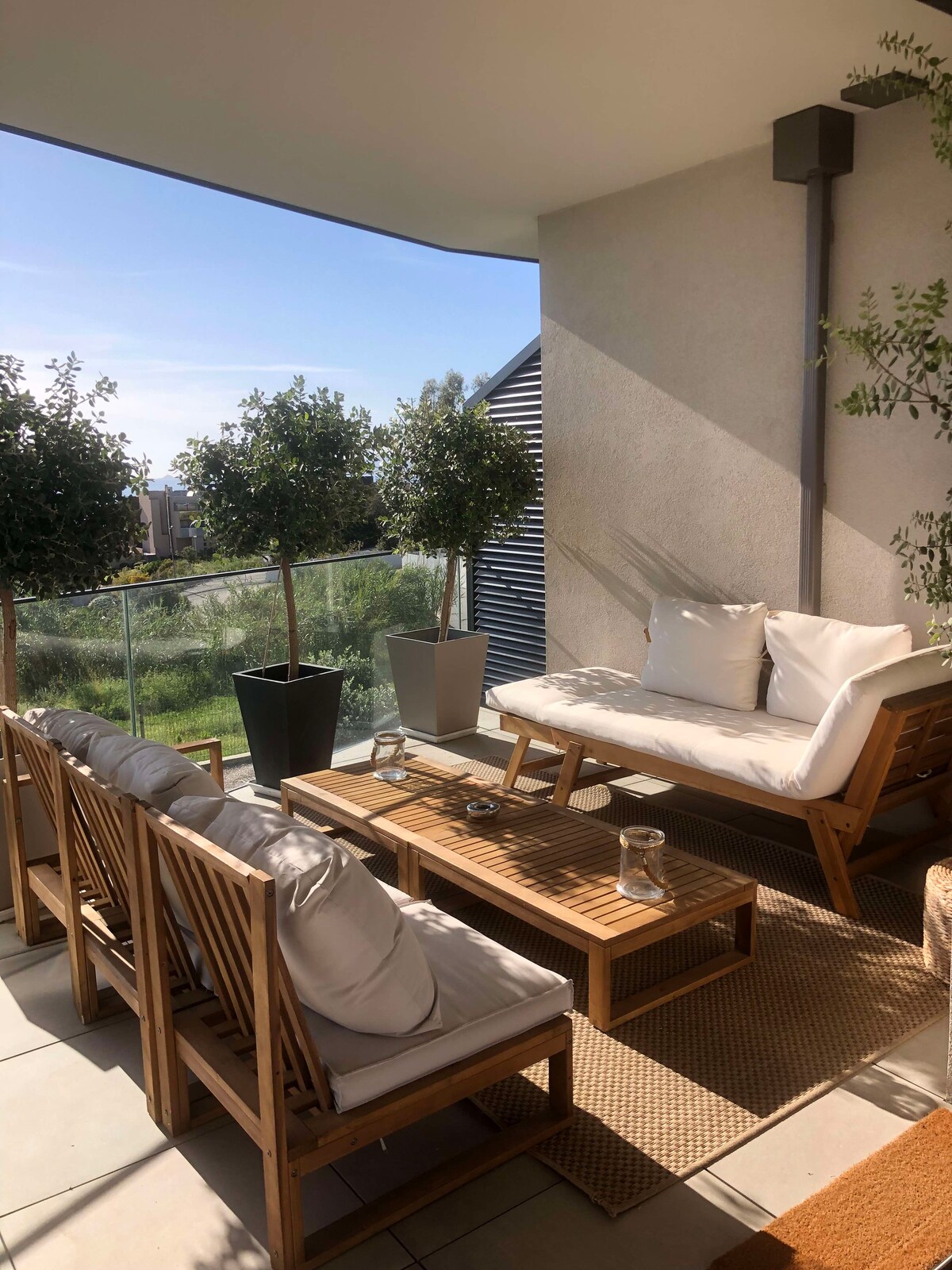 A terrace is featured with outdoor seating composed of wooden furniture and plush cushions. A low table made of wood is positioned in the center, surrounded by potted plants and a view of greenery in the background. Natural light illuminates the space.