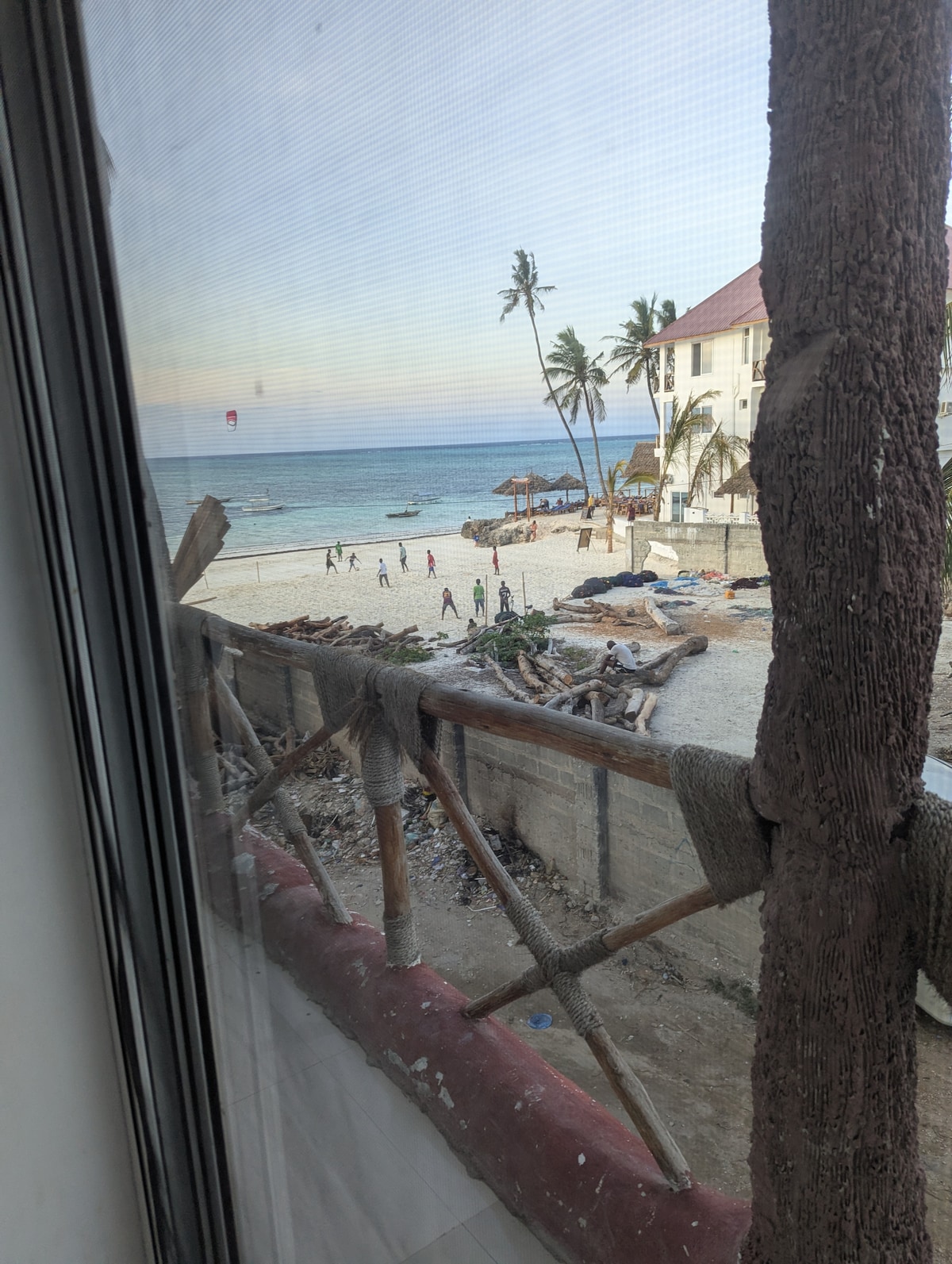 A view from a window reveals a beach scene with gentle waves lapping at the shore. Figures can be seen walking along the sandy beach, while palm trees sway in the breeze. Nearby, thatched umbrellas provide shade, and materials are scattered along the coastline.