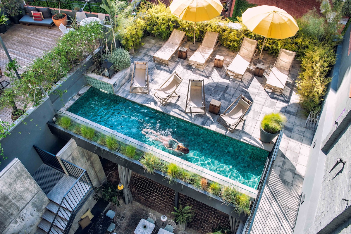 An inviting outdoor pool is surrounded by lounge chairs and large yellow umbrellas. Lush greenery frames the space, while a person swims effortlessly in the water. The area is designed to offer a relaxing retreat within the hotel grounds.
