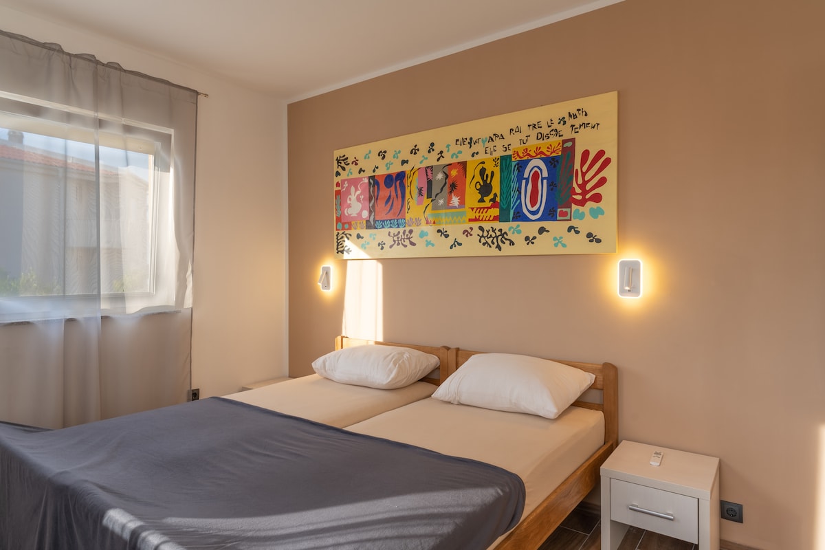 A bright sleeping area features a wooden bed frame with two spacious mattresses topped with white pillows. A colorful artwork hangs above the bed, adding a vibrant touch to the neutral wall. Natural light enters through a window adorned with sheer curtains.