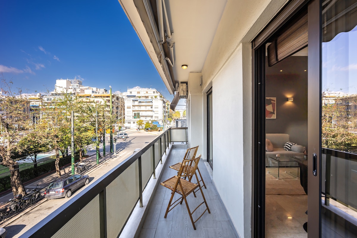 A private balcony is furnished with two wooden chairs, offering an open view of the vibrant street below. Natural light brightens the space, enhancing the modern design elements of the apartment. The peaceful setting invites relaxation while overlooking the lively atmosphere of Athens.