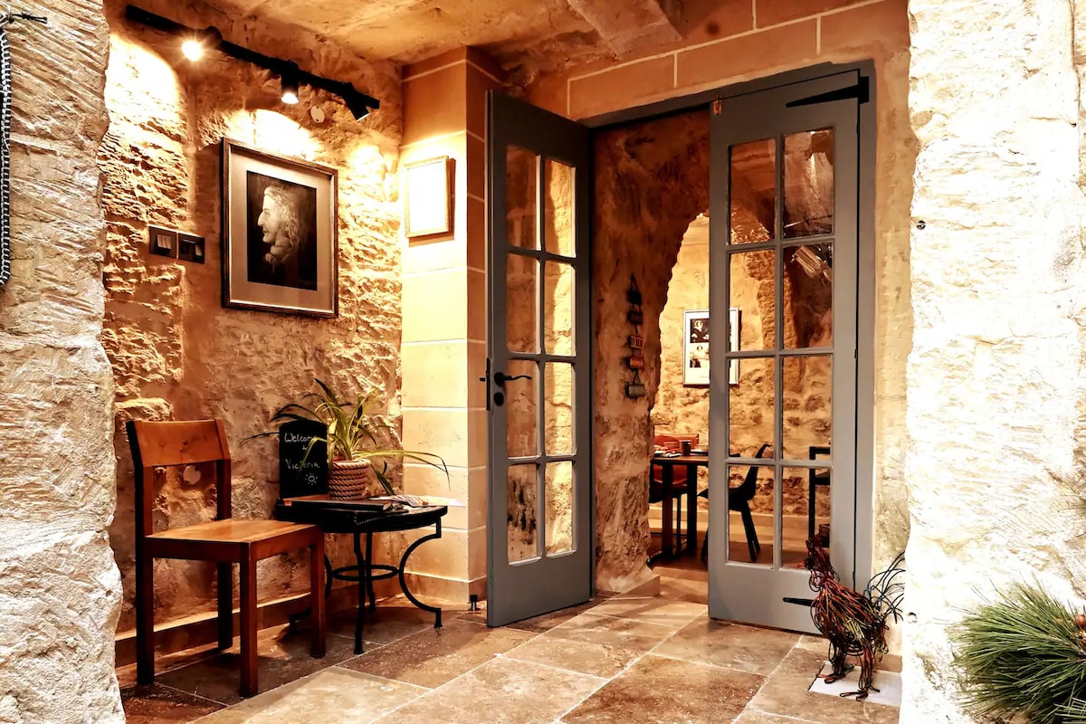 An inviting entrance features stone walls and a warm ambiance. Double doors with glass panes open to reveal a cozy dining area. A wooden chair and a small table are positioned on one side, enhancing the welcoming feel of the space.