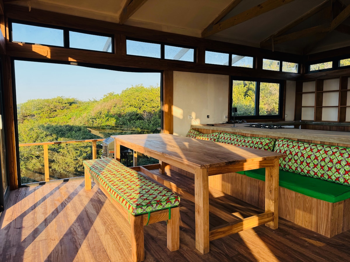 A spacious dining area features a large wooden table surrounded by a bench and a green cushioned seating area. Large windows allow natural light to illuminate the space, while views of the lush greenery can be seen outside.
