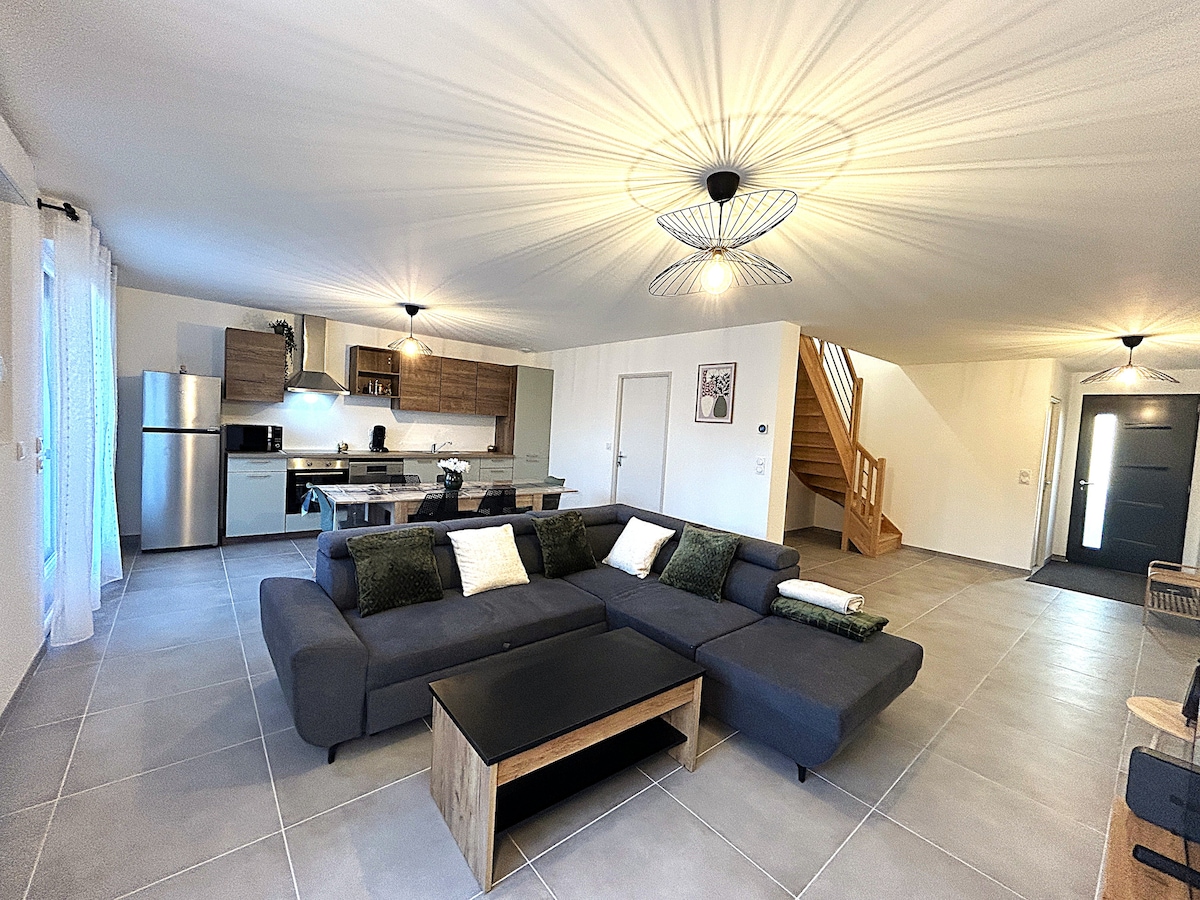 A spacious living area features a comfortable sectional sofa arranged around a modern coffee table. The open kitchen is visible, with wood cabinetry and appliances, while large windows allow natural light to fill the space. A staircase leads to the upper floor.
