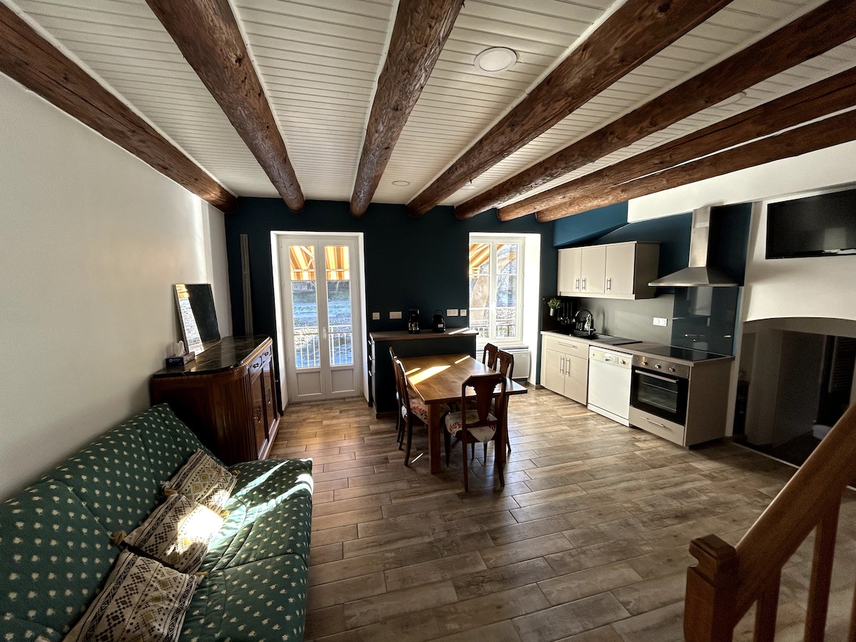 The spacious living area features exposed wooden beams and a large window allowing natural light to fill the room. A comfortable couch and a dining table with chairs create an inviting space. Modern kitchen appliances and cabinetry are present, enhancing functionality.