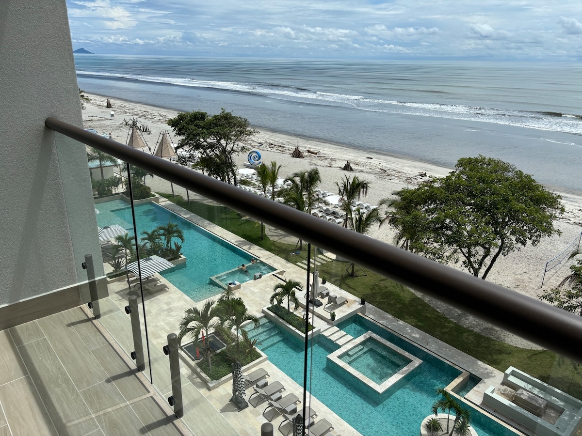 A spacious balcony view reveals the inviting pool area bordered by palm trees and lounges. In the distance, the sandy beach stretches along the coastline, leading to the tranquil sea with gentle waves. The wide sky is adorned with soft clouds, enhancing the serene atmosphere.